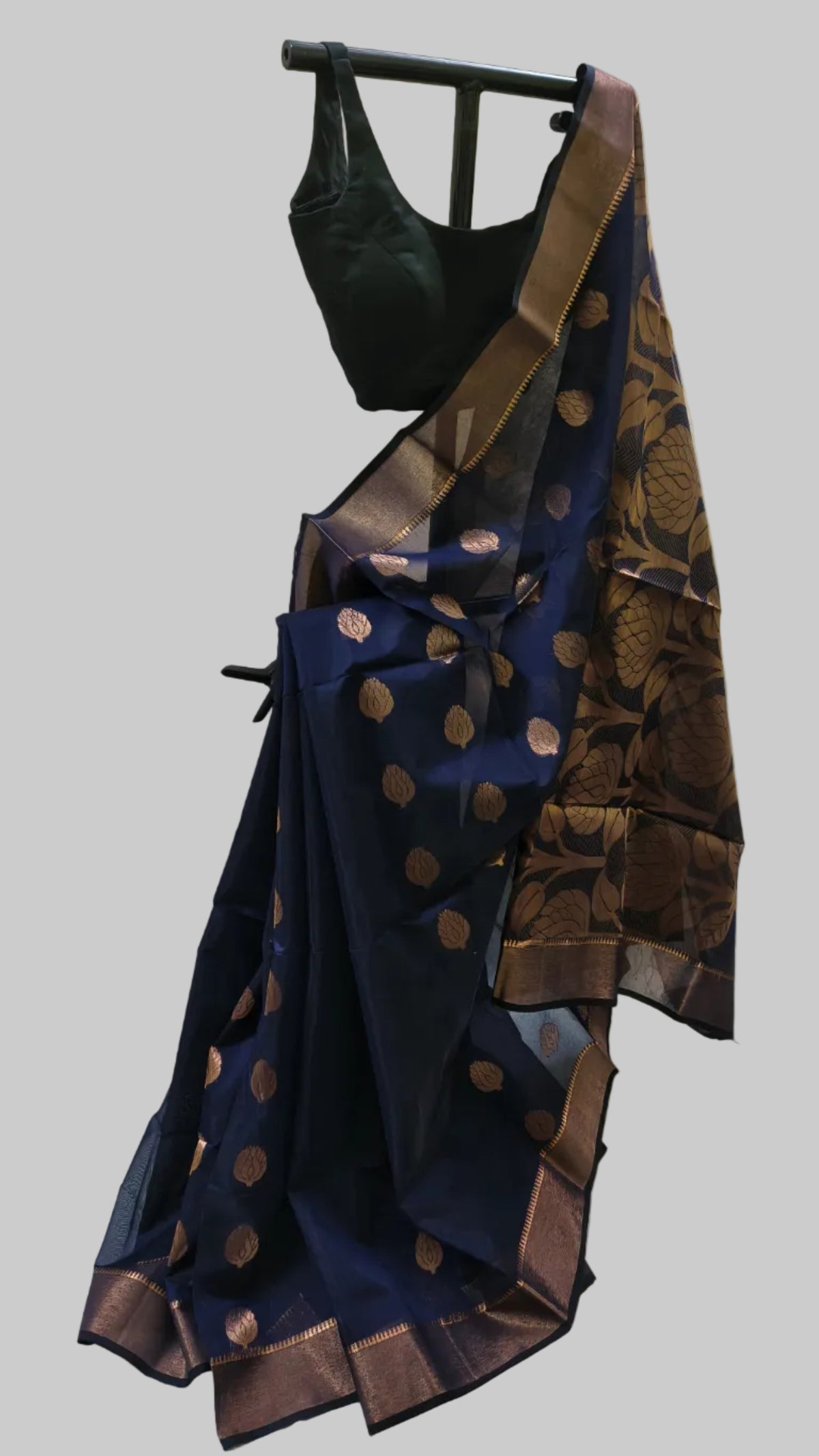 A navy blue silk saree with metallic bronze-gold zari motifs and border, paired with a black blouse, displayed on a black clothing hanger against a draped off-white fabric backdrop.