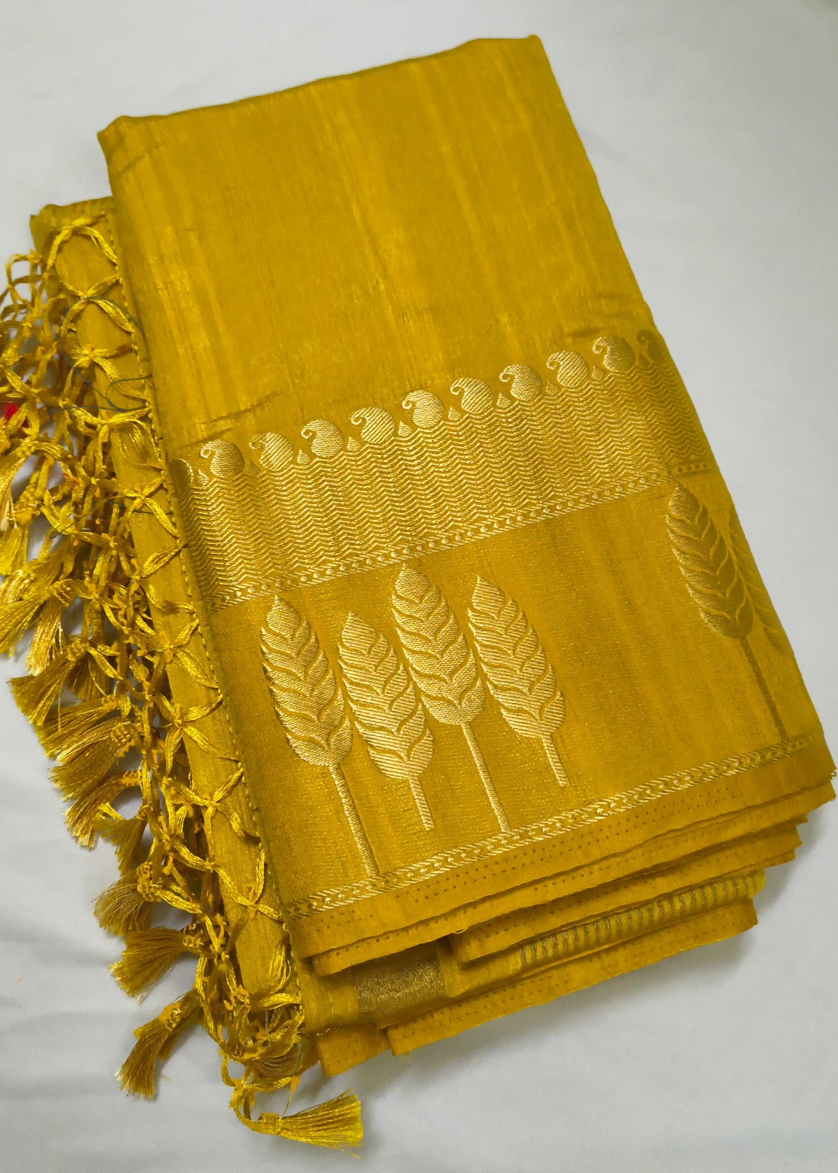 A folded mustard yellow silk saree or fabric piece with gold zari embroidery depicting wheat stalks and a decorative border, finished with a matching yellow tassel fringe on the left side, displayed on a white fabric background.