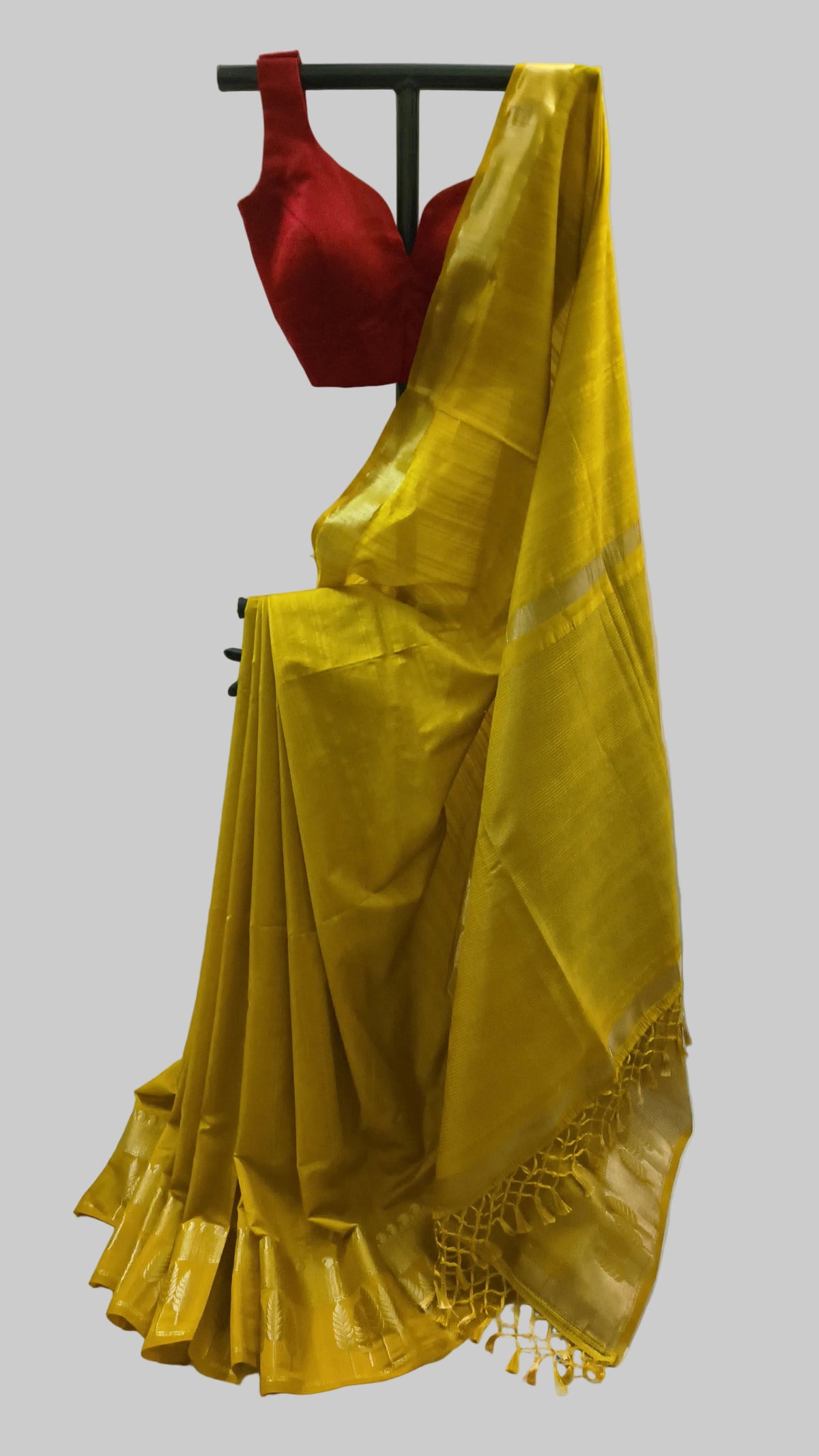 A yellow silk saree draped on a black hanger stand, paired with a red sleeveless blouse, set against a draped off-white fabric backdrop.