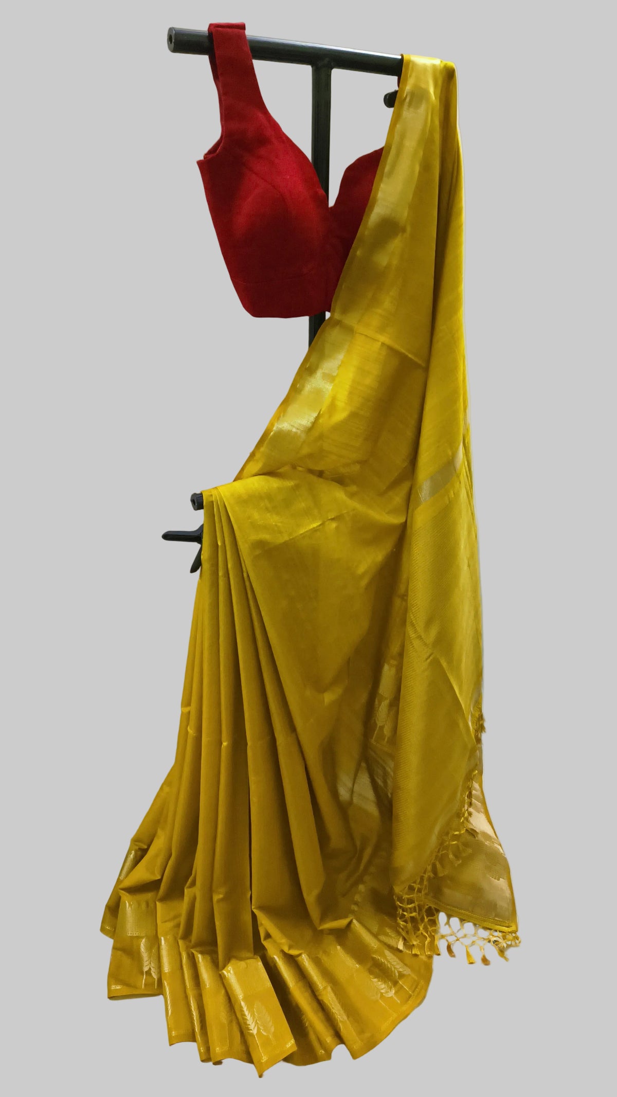 A yellow silk saree draped on a black clothing stand, paired with a red sleeveless blouse, set against a white fabric backdrop.