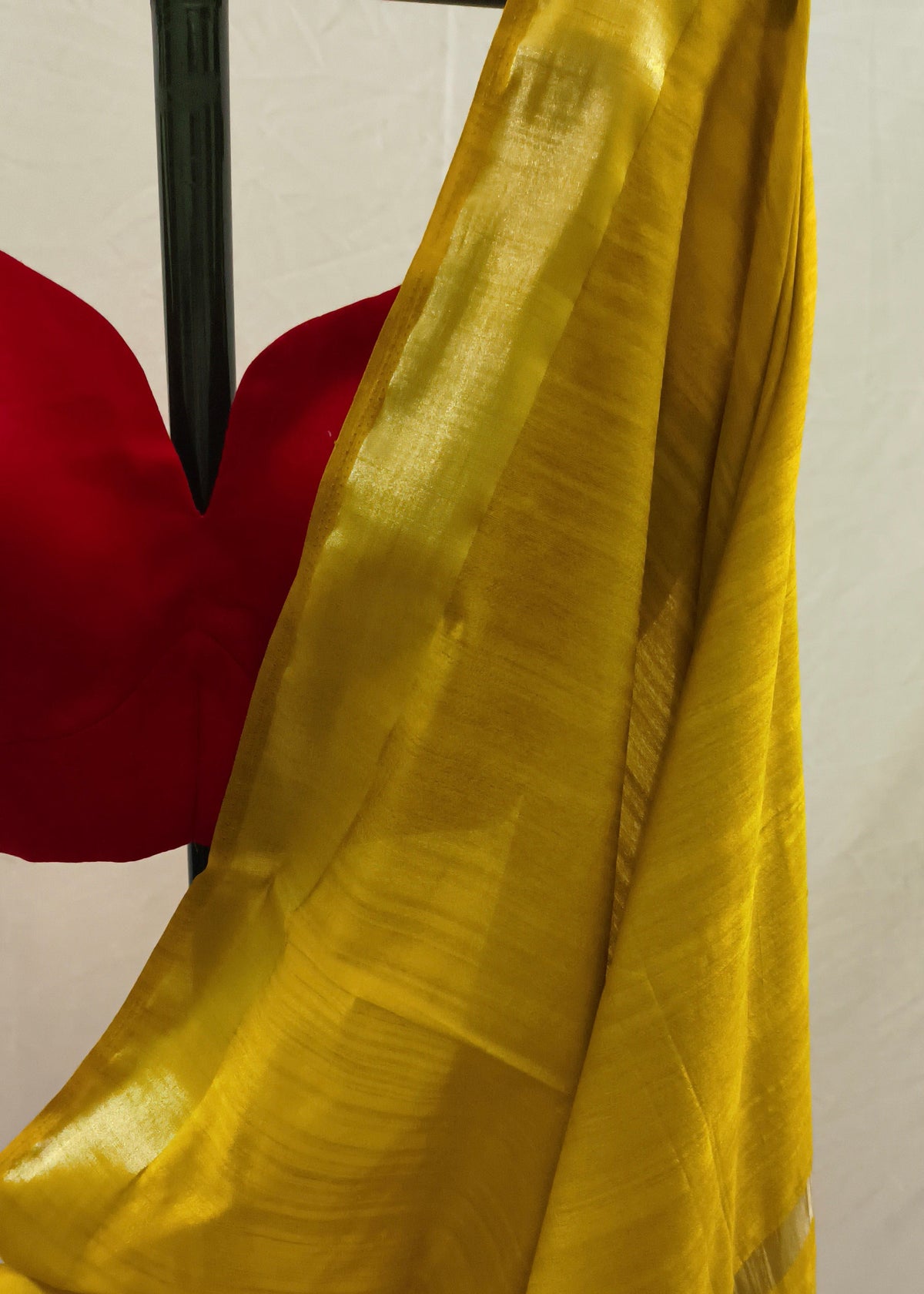 A close-up of a draped golden-yellow silk saree with a subtle sheen, paired with a red blouse, hanging on a black hanger against a light background.