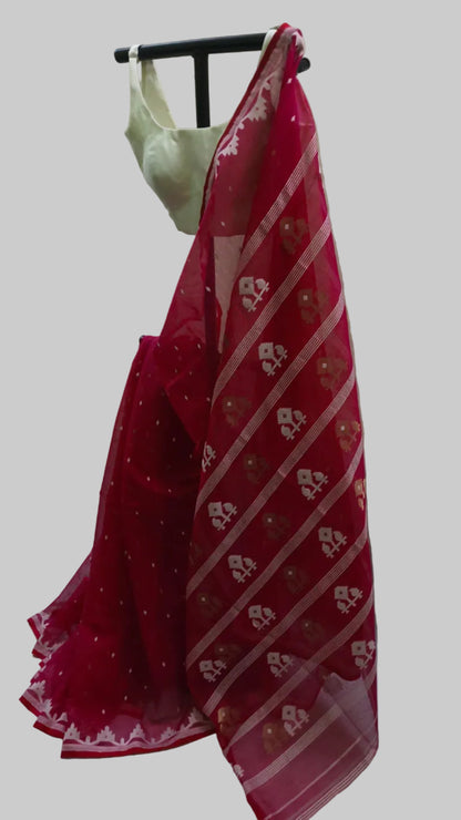 A red saree with white and gold woven patterns draped over a black hanger, paired with a light green blouse, set against a white fabric backdrop.