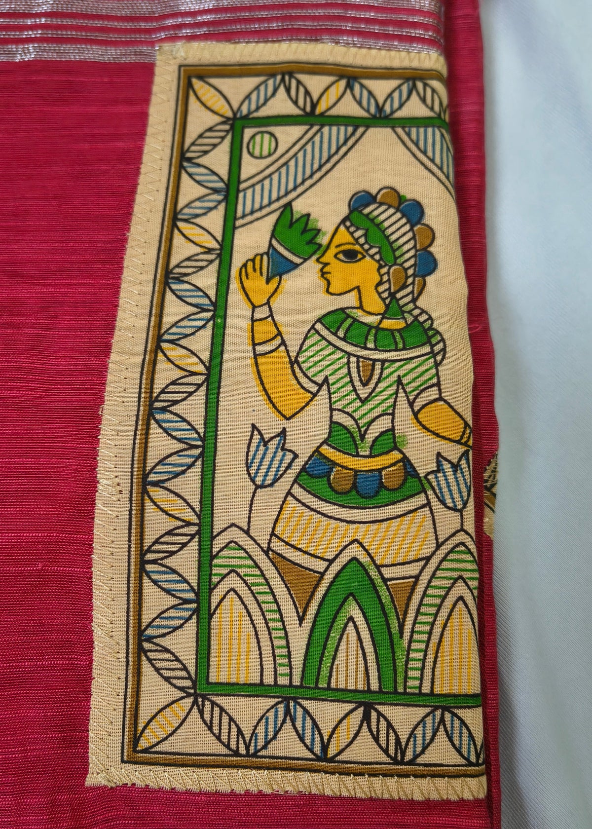 A close-up of a red and beige woven textile featuring a hand-painted style illustration of a woman in green, yellow, and blue within a rectangular border.