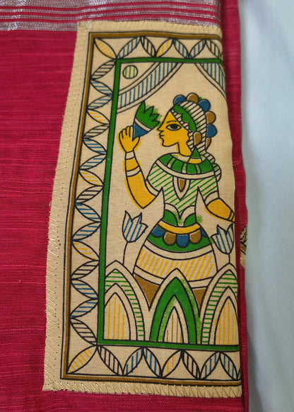 A close-up of a red and beige woven textile featuring a hand-painted style illustration of a woman in green, yellow, and blue within a rectangular border.