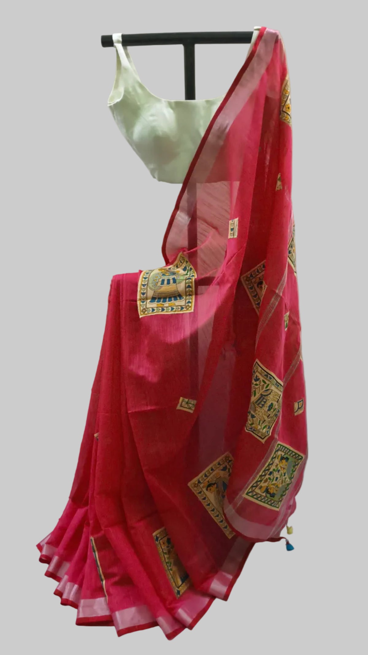 A bright pink saree with gold and green printed motifs draped on a black hanger, paired with a light green blouse, set against a draped white fabric backdrop.