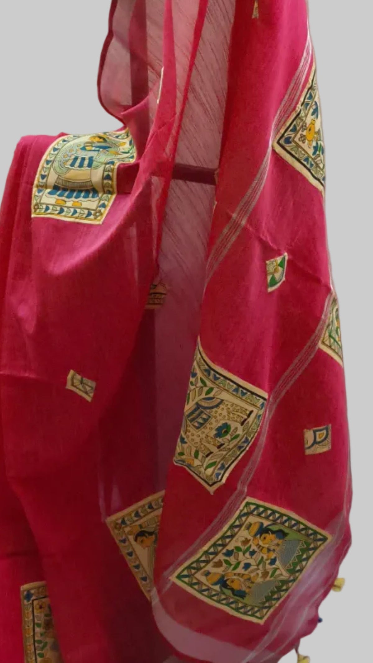 A bright pink saree with gold and green geometric patterns draped over a black hanger, paired with a light green sleeveless blouse, set against a draped off-white fabric backdrop.