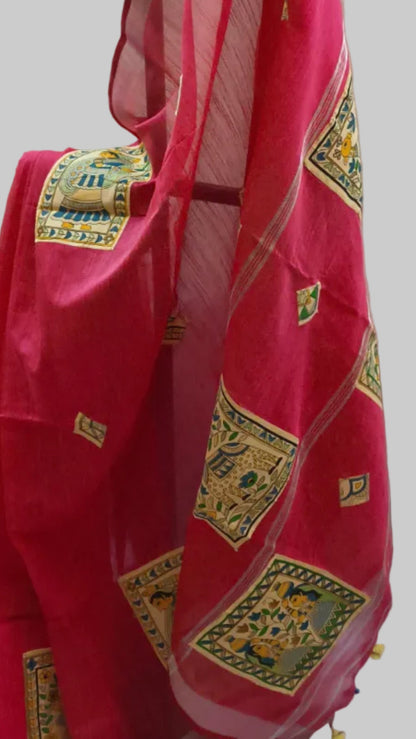 A bright pink saree with gold and green geometric patterns draped over a black hanger, paired with a light green sleeveless blouse, set against a draped off-white fabric backdrop.