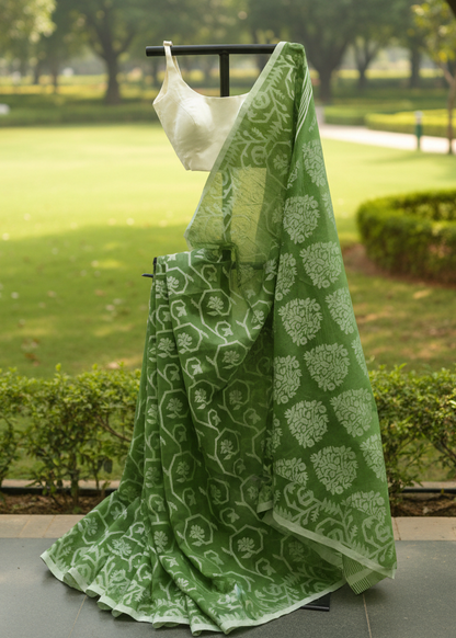 A green saree with white floral block prints is draped on a black hanger outdoors on a stone walkway, with a blurred park background featuring green grass, trees, and hedges.