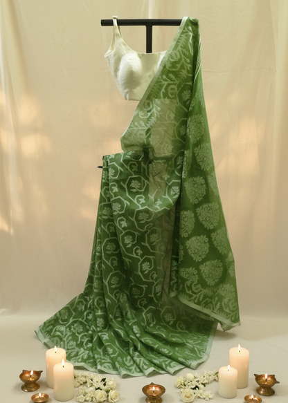 A green saree with a white floral block print pattern, draped elegantly on a black hanger stand. It is styled with a white sleeveless blouse, decorative white flowers, and lit candles on a neutral beige fabric backdrop.