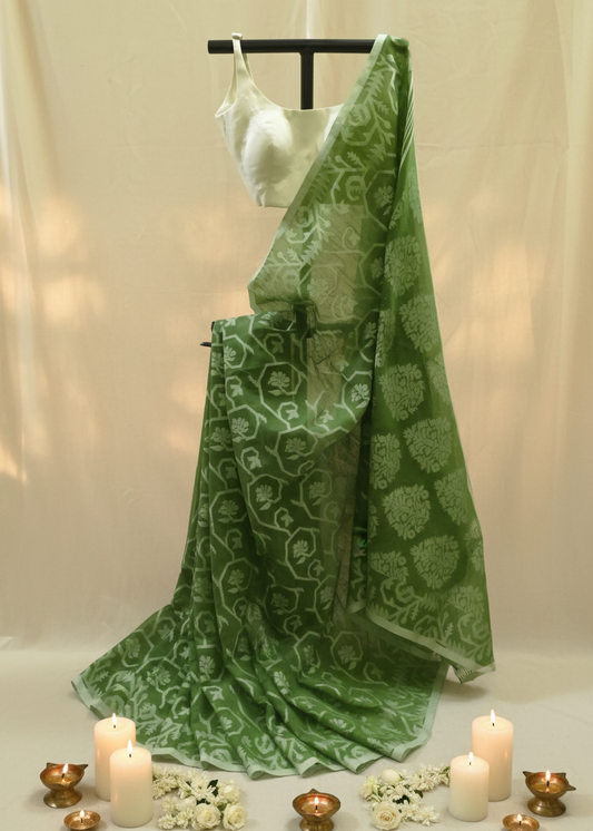 A green saree with a white floral block print pattern, draped elegantly on a black hanger stand. It is styled with a white sleeveless blouse, decorative white flowers, and lit candles on a neutral beige fabric backdrop.