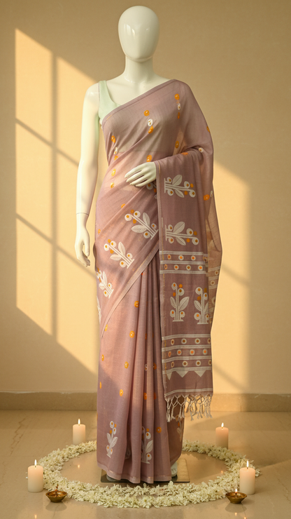 A mauve pink saree with white floral prints and gold accents, draped on a white mannequin. The background is a warm beige wall with window shadows, and the floor is decorated with white flowers and candles.