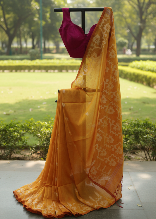 A yellow-orange saree with white floral patterns and a deep magenta blouse, displayed on a black clothing rack outdoors against a blurred green garden background.