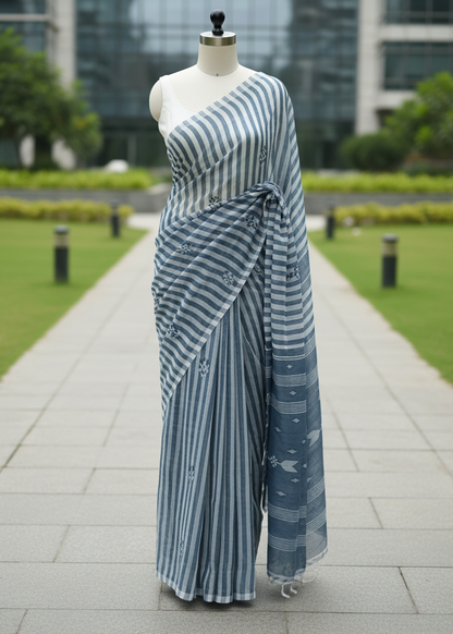 A blue and white striped saree draped on a mannequin torso, set against a blurred background of a modern glass building and green lawn.