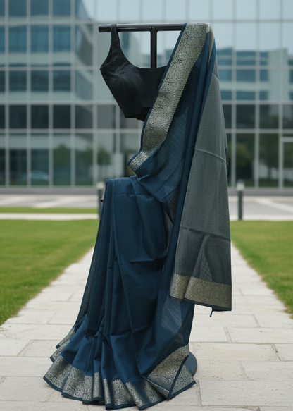 A dark teal blue saree with a wide silver border and a black sleeveless blouse, displayed on a black clothing hanger outdoors on a paved walkway with a modern glass building in the background.