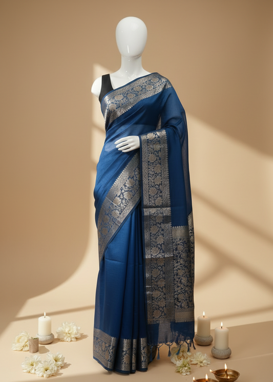 A royal blue saree with intricate silver and gold zari woven patterns, displayed on a white mannequin. The setting features a warm beige background with soft window shadows, decorative white flowers, and lit candles on the floor.