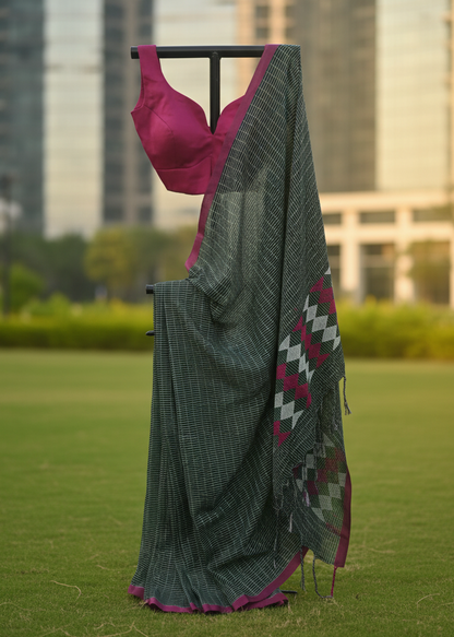 A dark gray and black checkered saree draped over a black clothing stand, paired with a magenta pink blouse, set against a blurred background of a green lawn and city buildings.