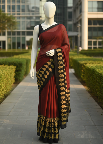A red and black saree with gold floral patterns displayed on a white mannequin outdoors on a paved walkway with green hedges and a modern building in the background.