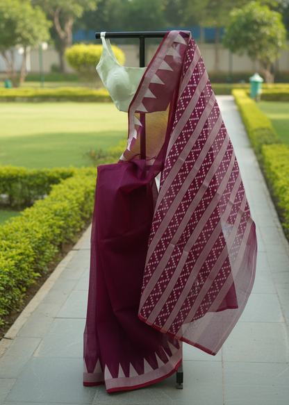 A deep magenta-purple saree with a white geometric patterned border and a white blouse draped on a black clothing rack, set against a blurred background of a garden pathway.