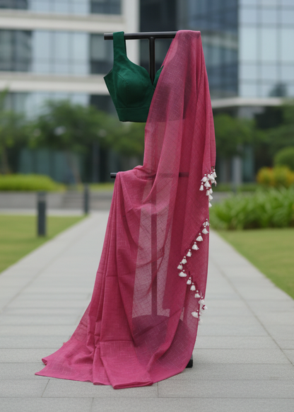 A pink sheer saree drape with white tassel trim hanging from a black clothing rack outdoors on a paved walkway, with a dark green blouse visible at the top and a modern glass building in the background.