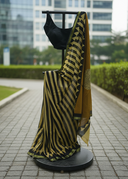 A black and mustard yellow striped saree draped on a black clothing display stand outdoors on a paved walkway, with a black blouse draped over the stand and a blurred modern building and green hedges in the background.